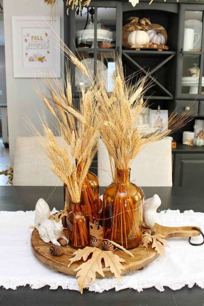 A table adorned with a tray of wheat and fall decorations, featuring a trio of amber apothecary vases.