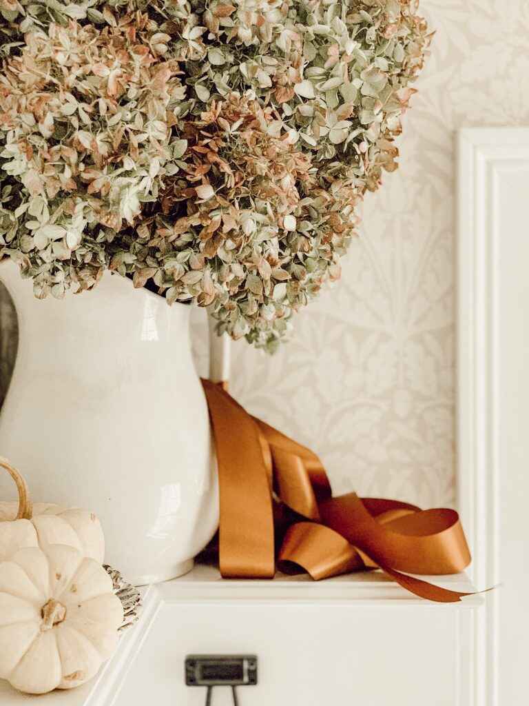 A vintage ironstone pitcher holds vibrant flowers beside white pumpkins, elegantly arranged on a wooden dresser.