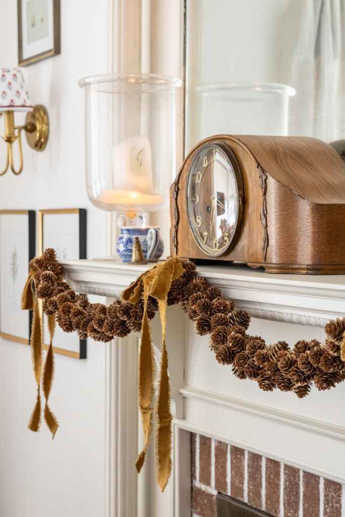 A vintage wooden clock sits on a mantle adorned with a gold ribbon garland and pinecones, creating a warm, inviting atmosphere.