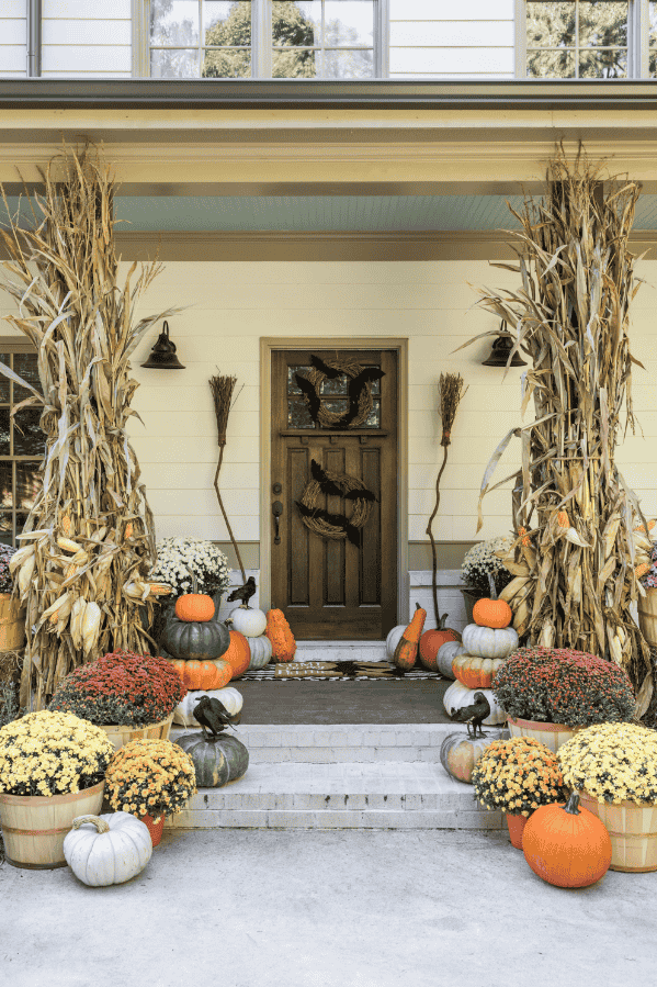 Charming front porch featuring pumpkins, corn stalks, witches' broom, and whimsical bats, perfect for Halloween spirit.